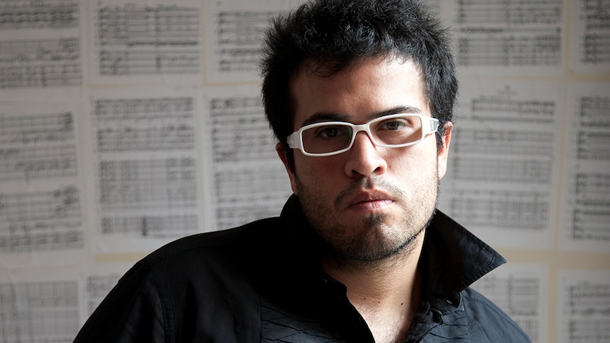 A photo of bandoneon player JP Jofre wearing white glasses. Blurred in the background is sheet music pasted on the wall.