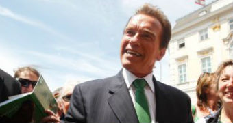 Arnold Schwarzenegger stands smiling in the street surrounded by a crowd.