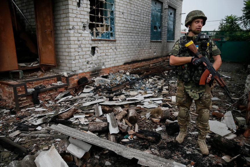 A man in army fatigues stands next to a destroyed building, with debris strewn across the ground