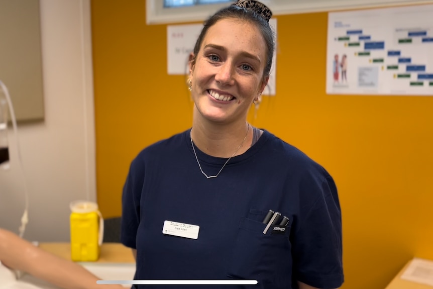 A young woman with her hair in a tight bun smiles at the camera, wearing a blue jumper, standing in front of a yellow wall.