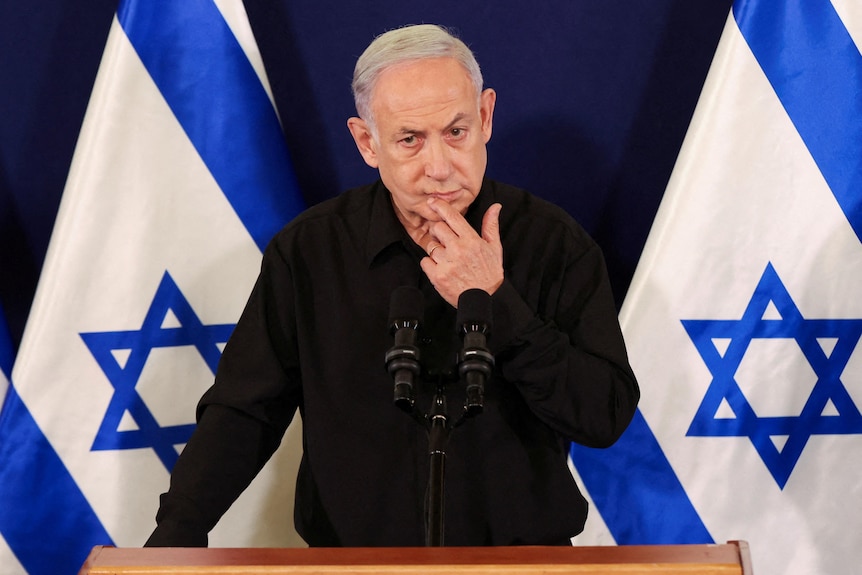 An older man with white hair puts his hand on his face, in front of two blue and white Israeli flags