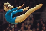 A young woman in blue leotard flies through the air with arms outstretched behind her, with blurred crowd behind her.