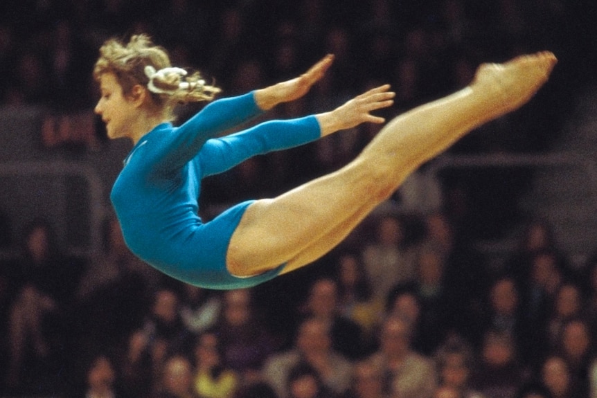 A young woman in blue leotard flies through the air with arms outstretched behind her, with blurred crowd behind her.