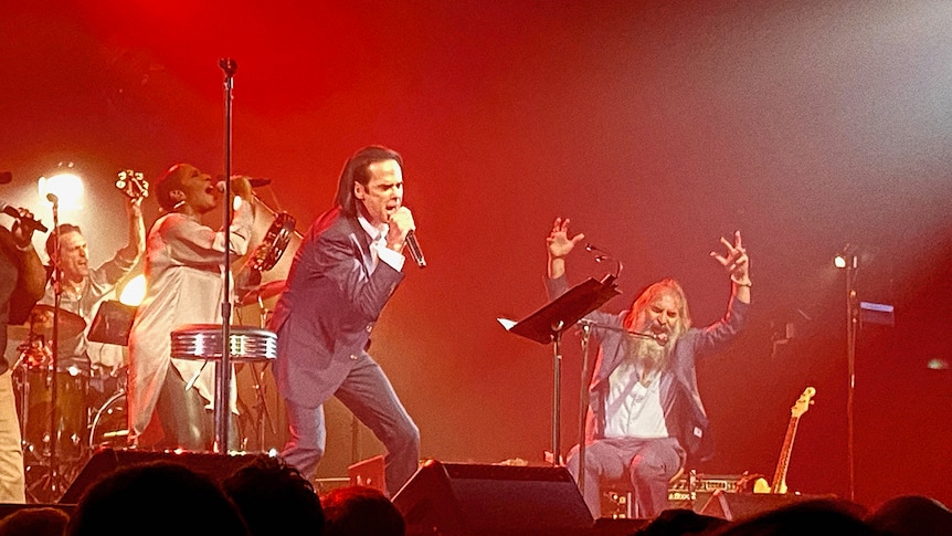 Nick Cave sings into a microphone, Warren Ellis throws his hands in the air, a backing vocalist and percussionist sing as well