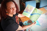 A woman with curly hair looking up at the camera and smiling, holding open a colourful booklet with watercolour drawings in it