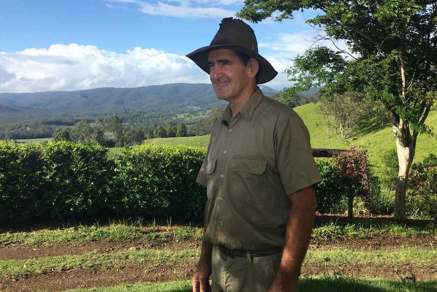 Man stands smiling on a farm, sunny day, very green hills and trees in the background.