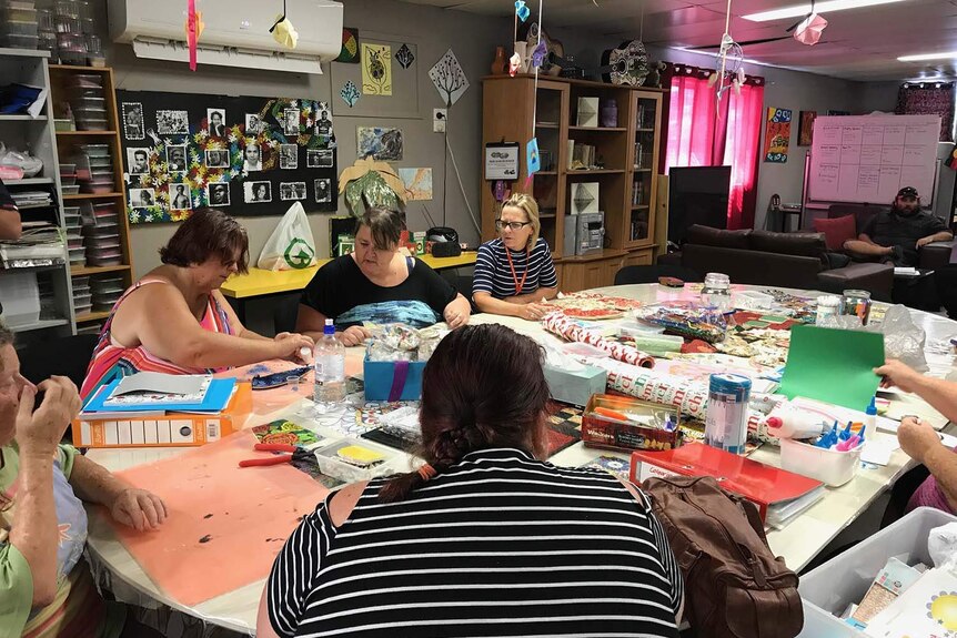 A group of women work on art projects around a large table while a man sits on an arm chair behind.