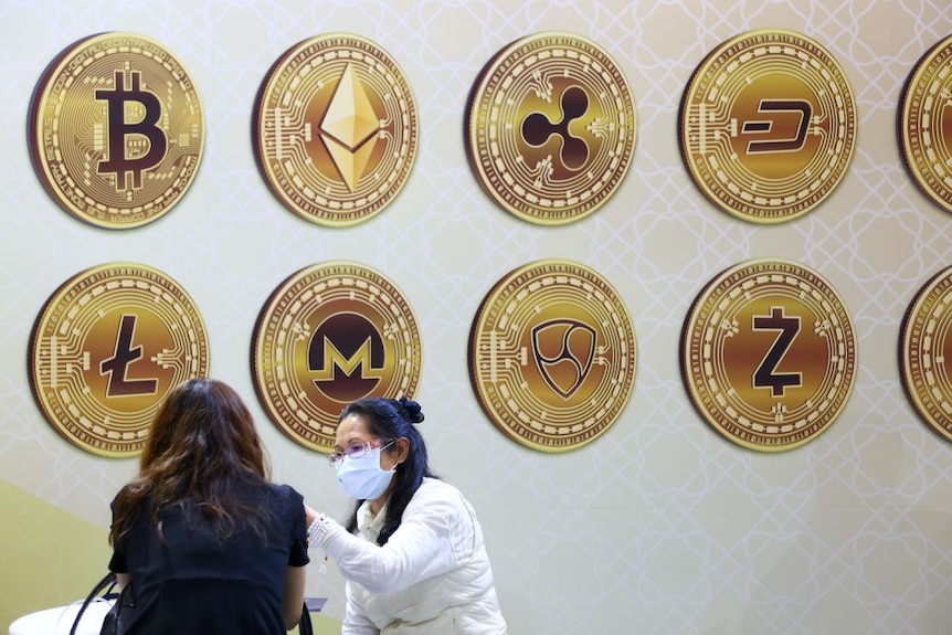 Two women talking, with large cryptocurrency logos being displayed on the wall behind them.