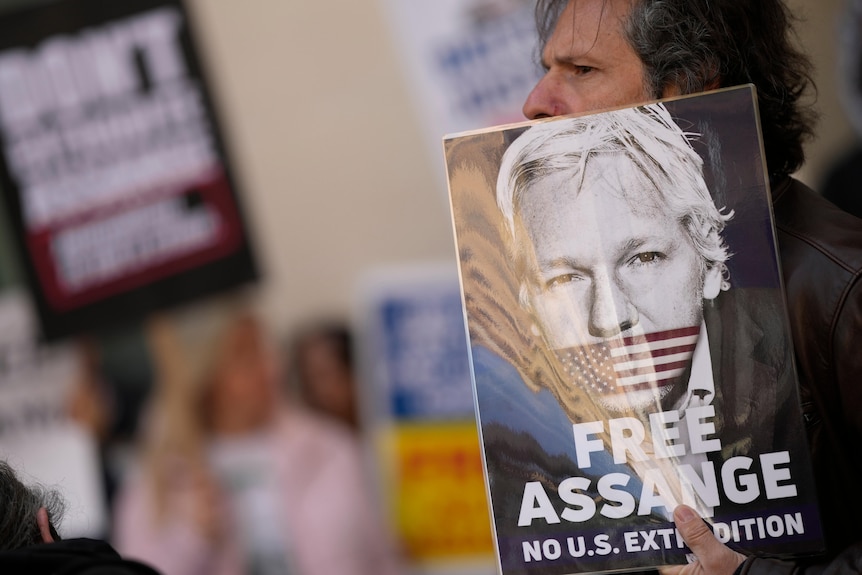 A man holds a poster over most of his face. The poster is of Julian Assange, and reads 'Free Assange'.