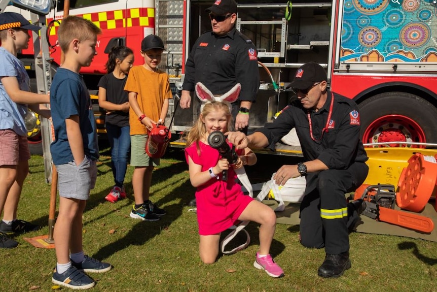 A young girl wearing bunny ears holds a fire hose while a firefighter helps guide her and other children surround a fire truck.