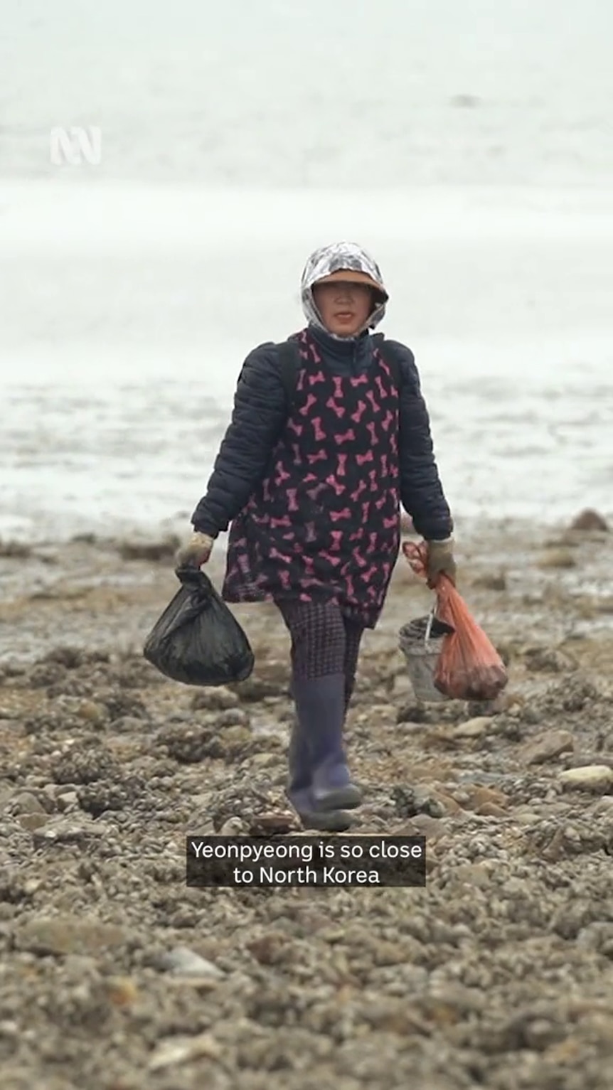 A woman with Asian features wearing gumboots walks on a rocky surface by the sea, plastic bags in each hand