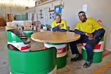 Two young men in tradesmen's clothes sit at a table made from upcycled oil drums.