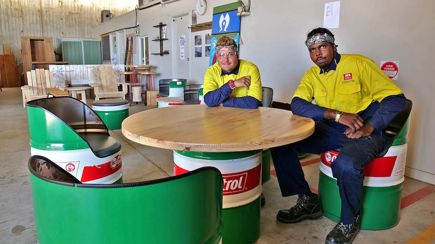 Two young men in tradesmen's clothes sit at a table made from upcycled oil drums.