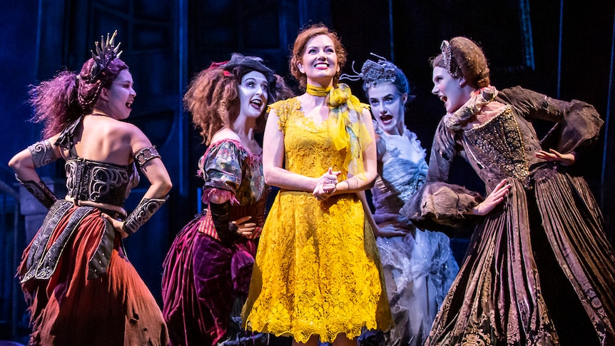 A woman in a bright yellow dress stands on stage, there are women surrounding her, dressed in dark colours. 