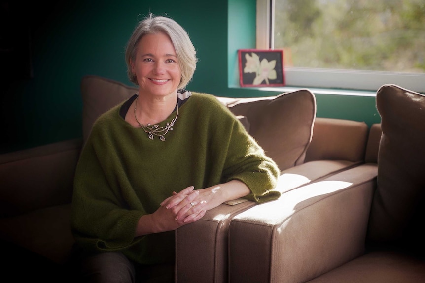 A woman with a sleek grey bob haircut, wearing a green jumper sits on a couch and smiles.