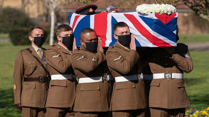 Soldiers in brown dress uniforms and face masks carry a coffin adorned with a UK flag, a military hat, a sword and a wreath.