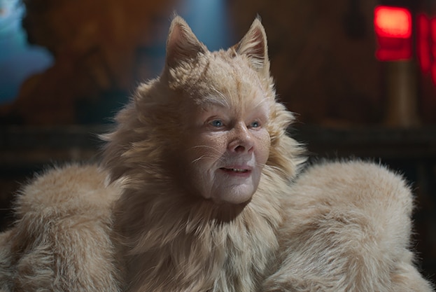 An oatmeal coloured CGI cat with a woman's face stands in a dark space, a small red light shines behind her.