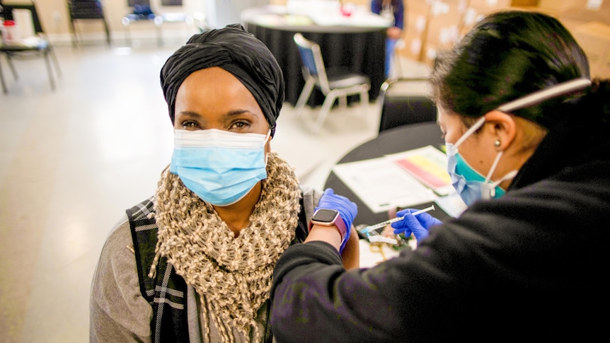 A woman in a blue face mask and black hair wrap sits while a nurse injects a needle into her shoulder