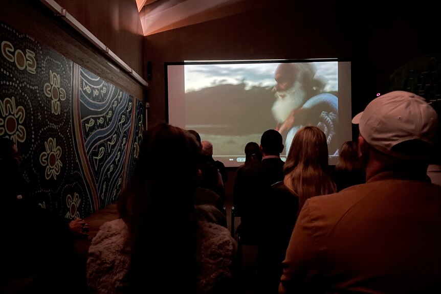 People sitting watching a film next to an Idigenous painting on the wall.