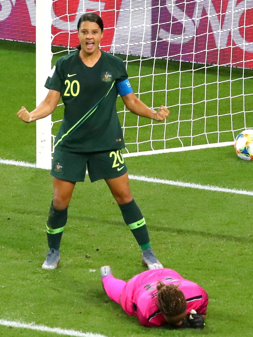 Sam Kerr stands with her arms taught in celebration. The Jamaican goalkeeper lies face down on the grass