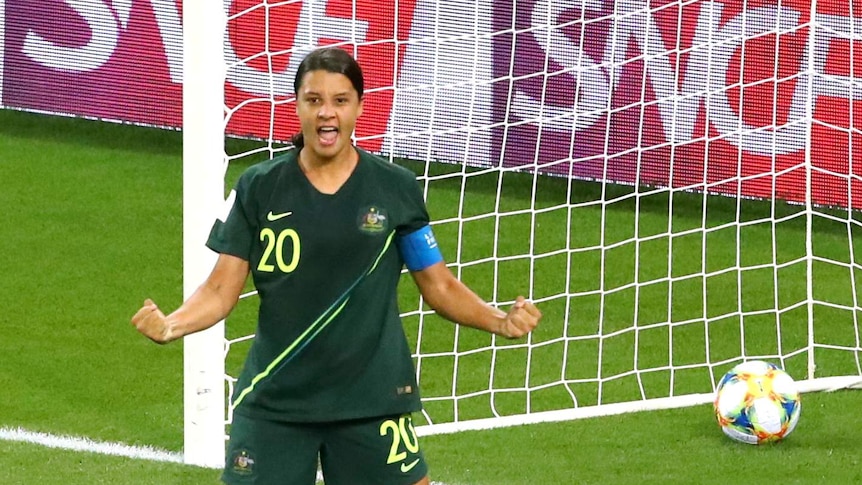 Sam Kerr stands with her arms taught in celebration. The Jamaican goalkeeper lies face down on the grass