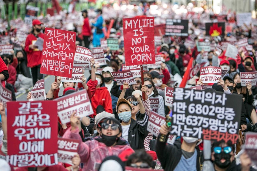 South Korean people protesting in the street; one holds a sign in English saying 'my life is not your porn'.