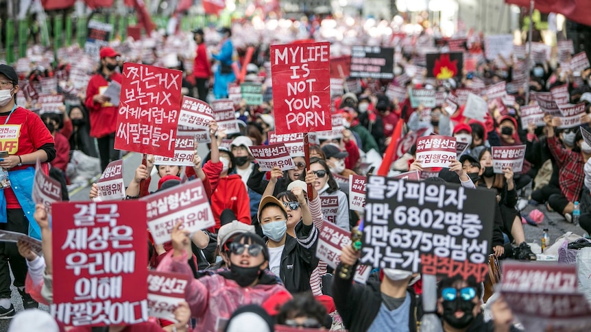 South Korean people protesting in the street; one holds a sign in English saying 'my life is not your porn'.