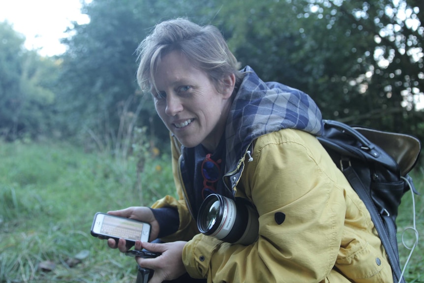 Poppy Stockell holding camera and phone crouched in forest in northern France.