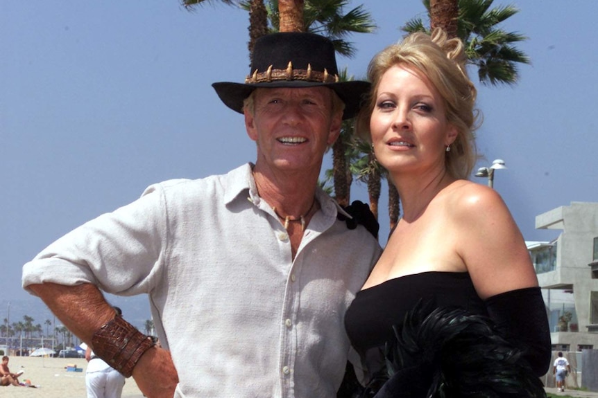 Paul Hogan and Linda Kozlowski stand in front of palm trees on a beach.