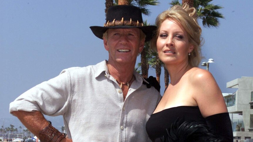 Paul Hogan and Linda Kozlowski stand in front of palm trees on a beach.