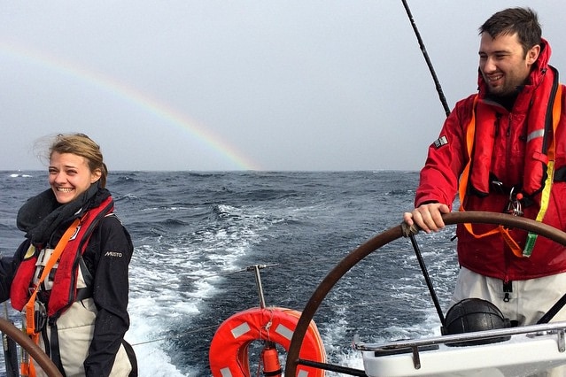 Jessica smiling, Cameron at the helm of a boat, ocean and rainbow behind.