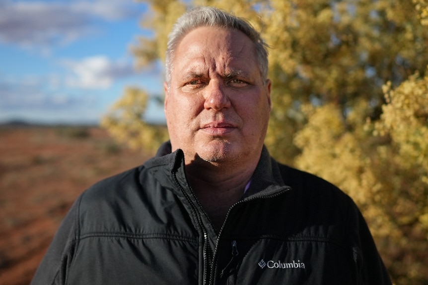 Grey haired man stands in front of a yellow tree and red-earthed paddock.