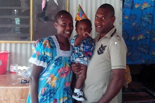 A ni-Vanuatu man in his 30s, holding his toddler son who wearing a party hat, and standing next to his wife. 