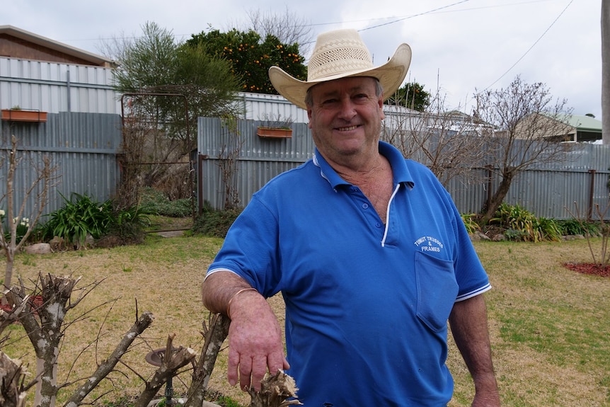 A middle-aged man wearing a straw hat smiles at the camera in his backyard while leaning on a bush
