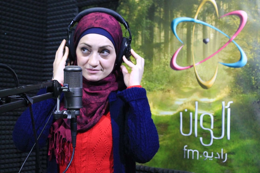 A woman in a scarf touches her headphones as she speaks into a microphone.