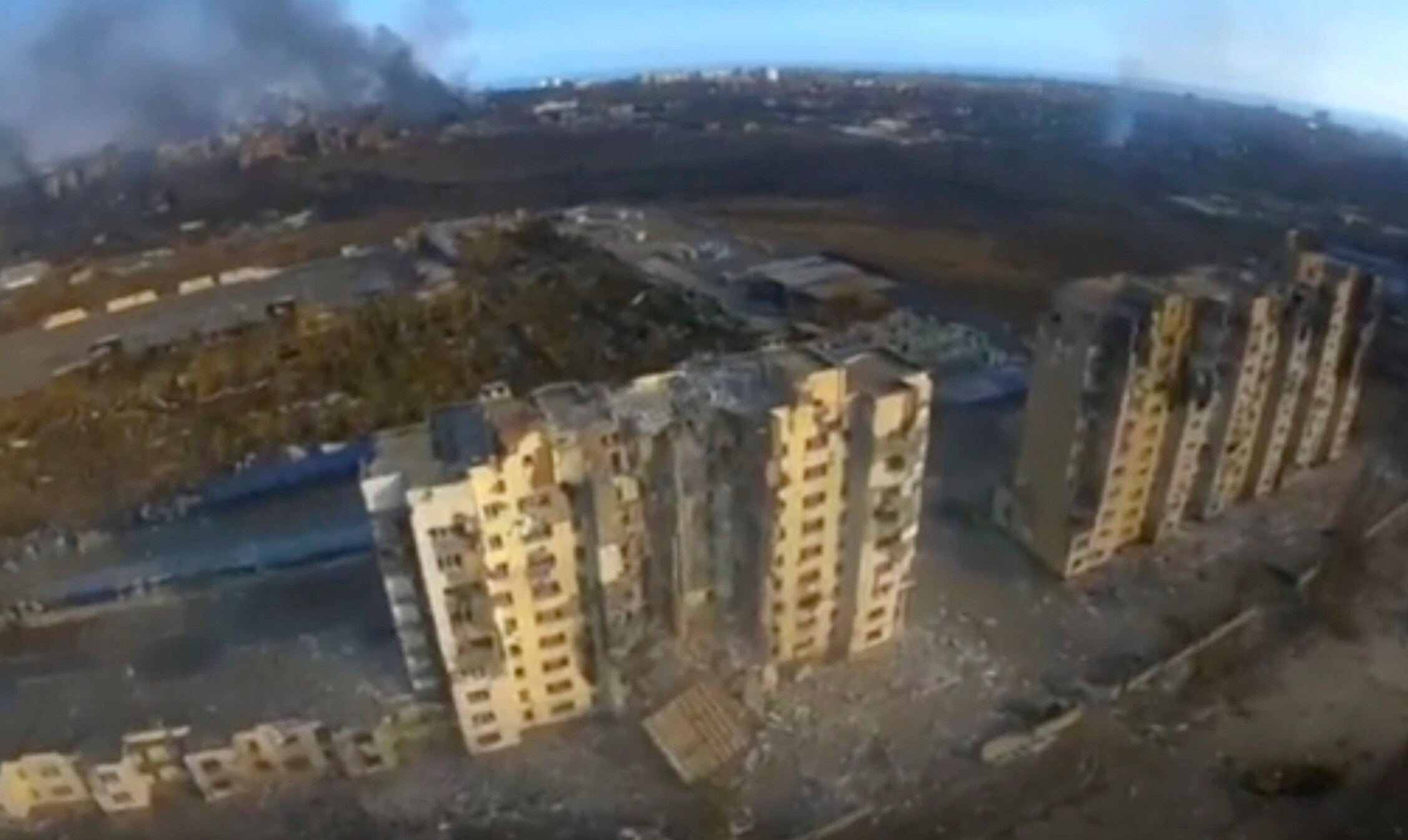 An aerial view of destroyed high-rise buildings, with smoke in the background.