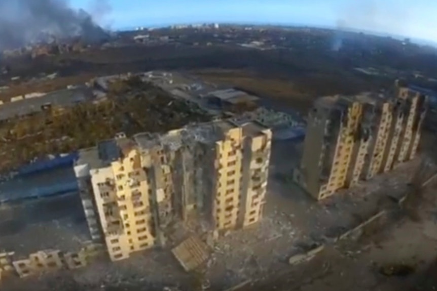 An aerial view of destroyed high-rise buildings, with smoke in the background.