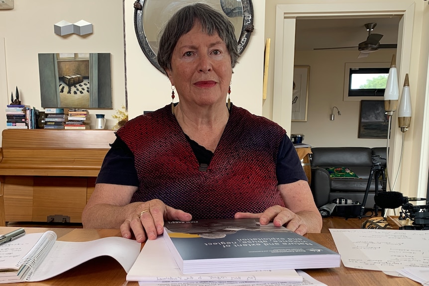 A middle-aged white woman sitting at a kitchen table. She is wearing a red blouse and has various paperwork in front of her