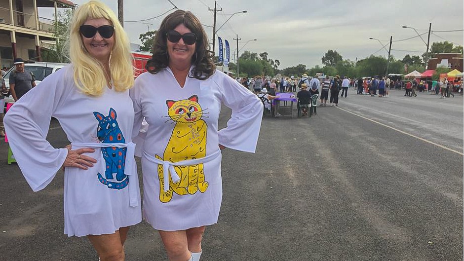 Two women in white 1970s dresses with animal prints on them stand in front of a wide country street