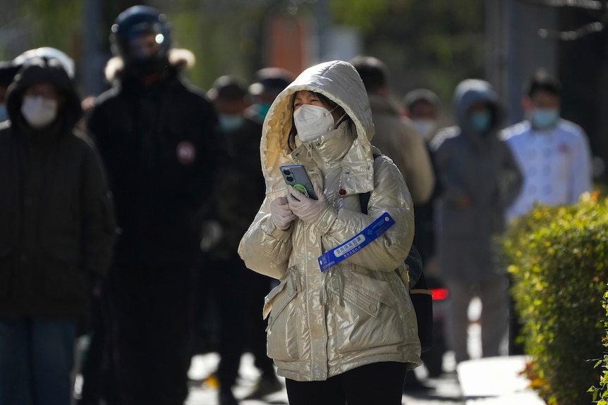 A woman wearing a winter coat, and a surgical mask over her face, looks up as she holds her phone to her chest