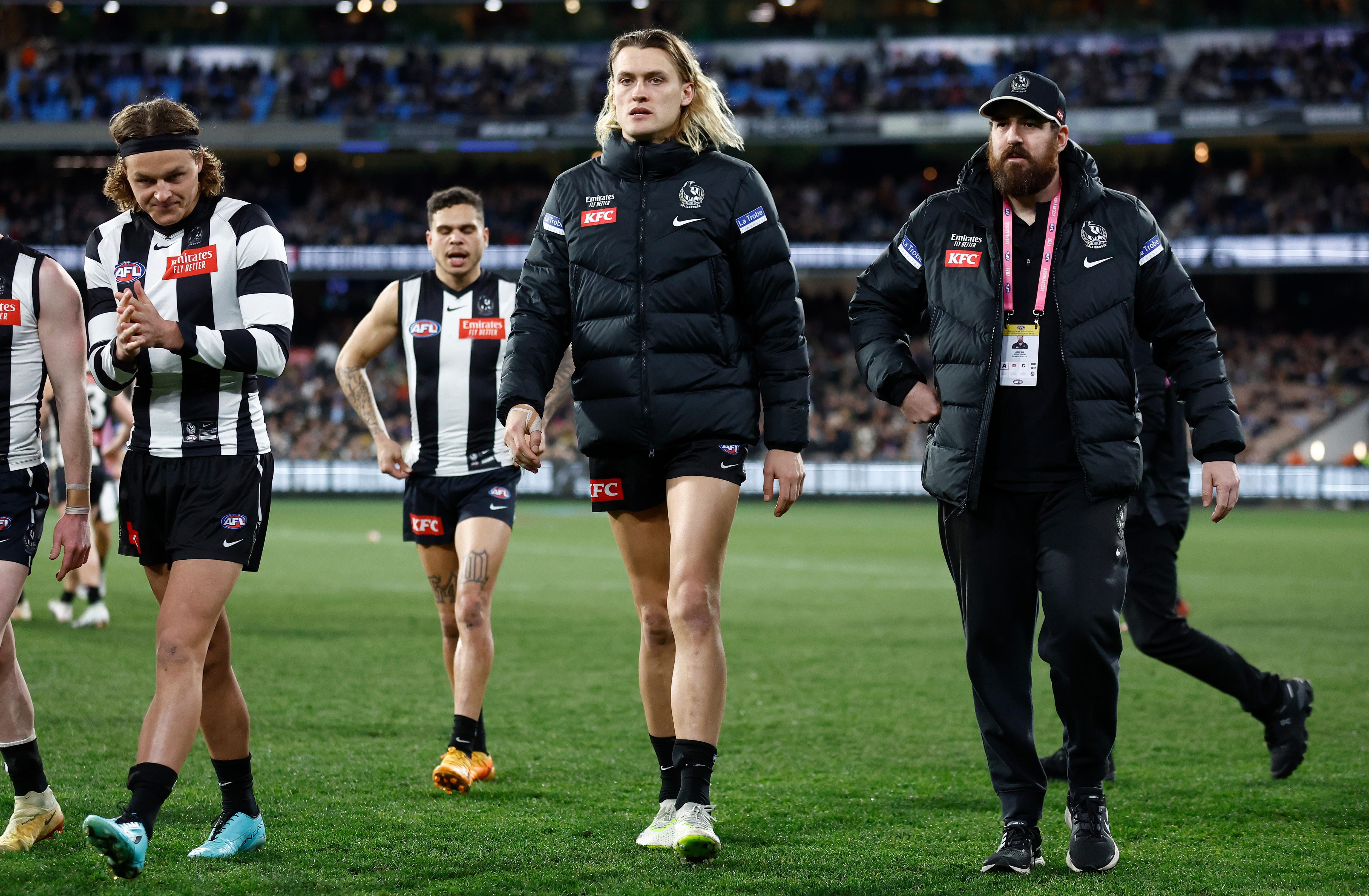 A Collingwood AFL captain walks off the ground with teammates and officials wearing a club jacket after being subbed.
