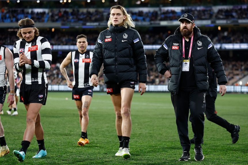 A Collingwood AFL captain walks off the ground with teammates and officials wearing a club jacket after being subbed.