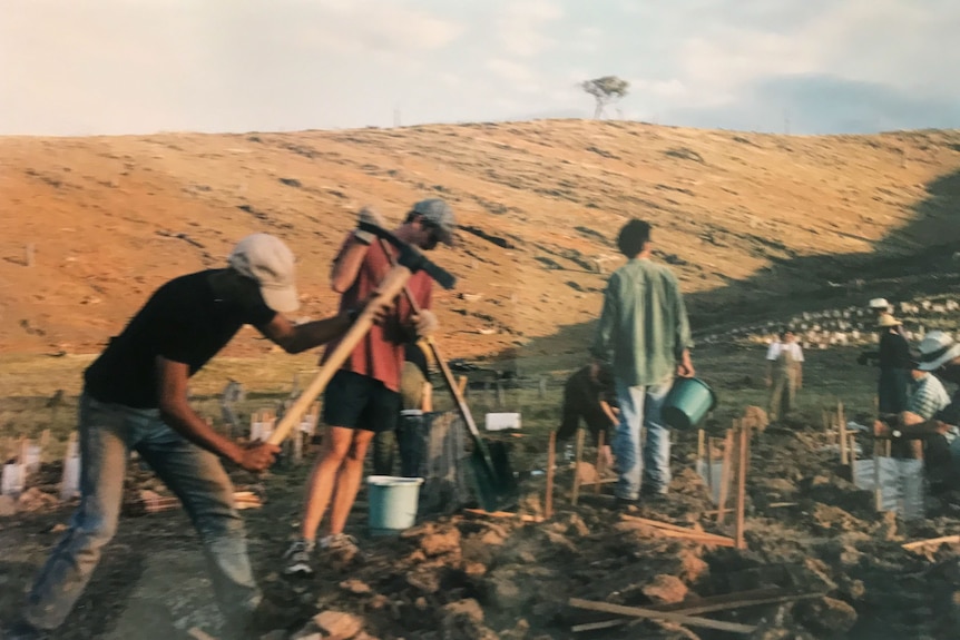 People work with picks and shovels in a bare paddock, behind them is the deforested hill