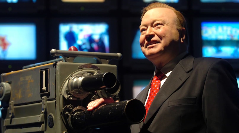 Television icon Bert Newton stands next to a vintage TV camera with a wall of modern TV screens behind him