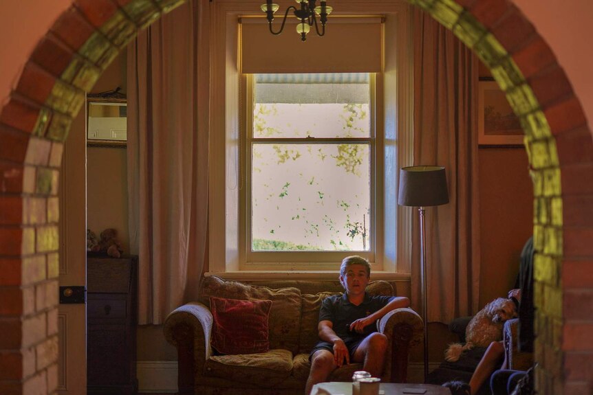 A teenage boys in school uniform sits on a couch comfortably in a dimly-lit living room.
