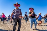 Two people wearing pink flamingo sunglasses dance while laughing and smiling in a crowd of people dancing in a dusty landscape.