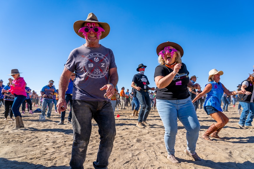 Two people wearing pink flamingo sunglasses dance while laughing and smiling in a crowd of people dancing in a dusty landscape.