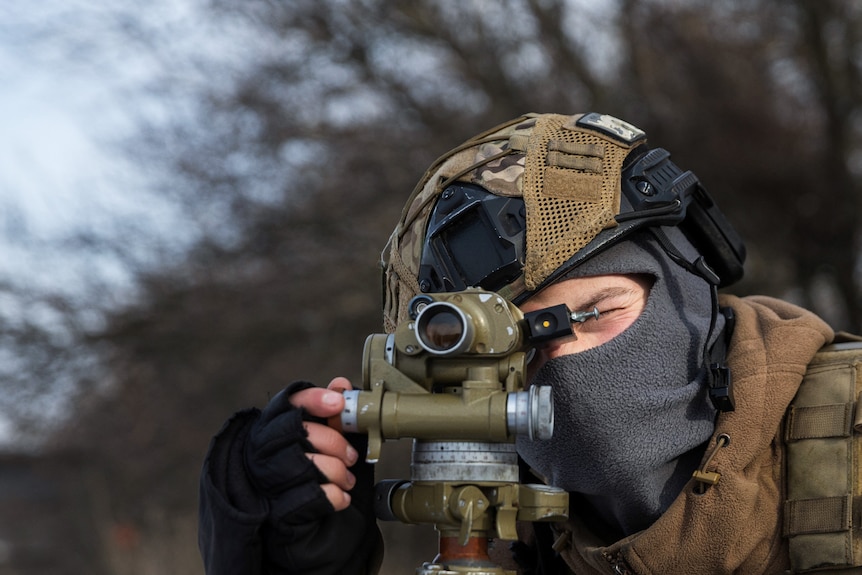A Ukrainian serviceman dressed in a helmet and wearing a grey mask looks through a rangefinder 