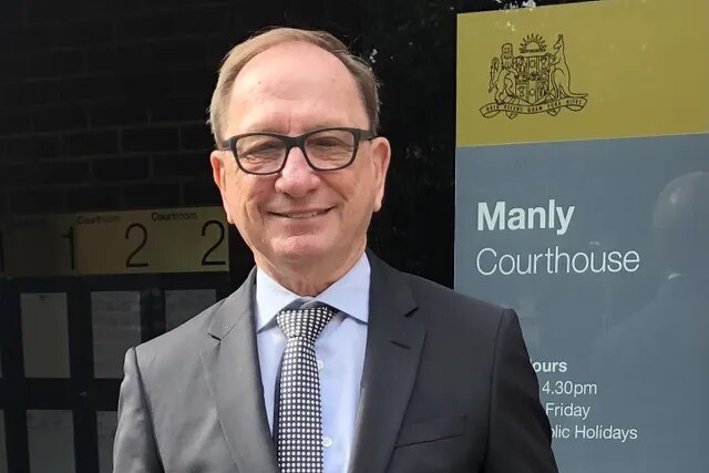 A smiling, bespectacled man of middle age, wearing a suit and standing outside a court building.