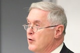 Robert Cribb wearing suit, blue time and silver rim glasses stands with grey wall in background.